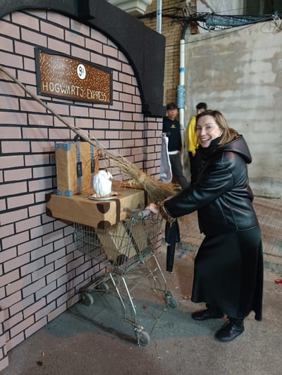The image depicts an outdoor, nighttime scene in El Port De Sagunt, Spain, centered on a Harry Potter-themed photo opportunity. A Caucasian woman, appearing middle-aged, stands on the right, smiling widely and looking towards the camera. She wears a black hooded jacket with leopard-print cuff accents, a long black skirt, and dark boots. Her right hand rests on the handle of a metal shopping cart. The shopping cart is loaded with prop luggage, including two brown, box-like suitcases with dark corner protectors; one is inside the cart, and a larger one is stacked on top. On the upper suitcase, a white owl figure sits within a thin wire cage, accompanied by a long, slender broomstick leaning against the luggage and a prop brick wall. This wall, painted light pink with dark mortar lines and featuring a dark arched top, displays a brown sign with white text reading "9 3/4 HOGWARTS EXPRESS." In the blurred background, behind the woman and the brick wall, two young males are partially visible; one in a yellow hoodie and another in a dark hoodie. The ground is paved, and an actual building wall with utility pipes and wiring is visible to the right of the prop wall.