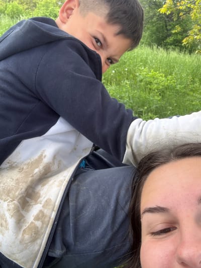 A photograph shows a woman and a boy outdoors on a grassy area. The woman is cropped from the top of the frame, showing only her eye and forehead. The boy is visible from the left side, looking mischievously at the camera with one eye partially obscured by his navy blue hooded sweatshirt. His sweatshirt has white interior lining stained with mud. The background consists of green grass and foliage. The location is possibly Liava Reka, Bulgaria.