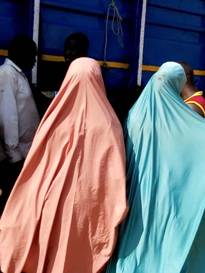 The image shows three individuals in the city of Gadao, Nigeria. Two individuals are wearing head coverings that obscure their faces and upper bodies, one in a peach-colored garment and the other in a light blue garment. A third individual, dressed in a white shirt, is partially visible on the left. The background consists of a blue structure with yellow and white horizontal accents, likely part of a vehicle or building. The scene appears to be outdoors, possibly in a market or transportation hub.