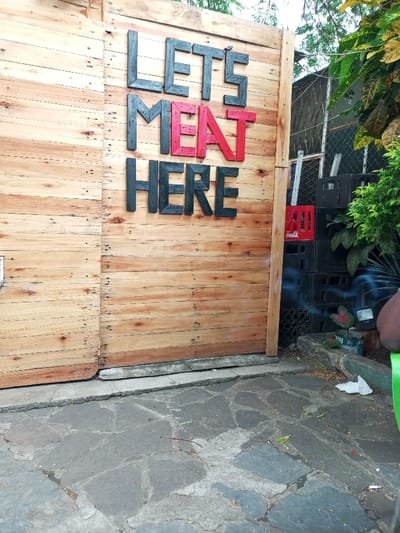 The image shows a wooden wall with the words "LETS MEAT HERE" written in black and red letters, located at the coordinates -25.976, 32.591 in Maputo, Mozambique.  There are stacked crates to the right of the wooden wall. To the right of the crates is a small patch of grass with a few green leaves.  The ground in front of the wall is made of small, gray, uneven stones.  A portion of a person's leg and the bottom of a green chair can be seen in the bottom right corner.  The scene appears to be in front of a restaurant or eatery.