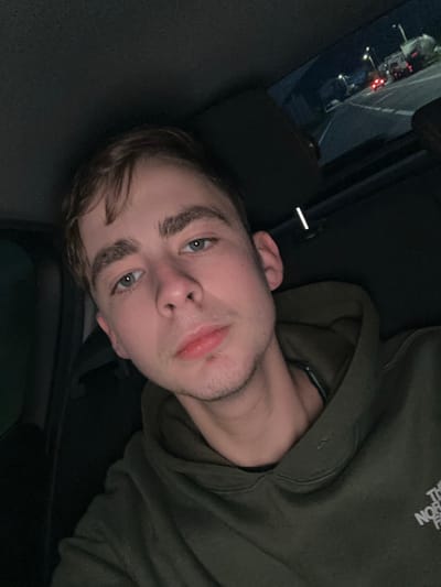A young man lies down inside a vehicle, wearing a green hoodie. He gazes upwards with light-colored eyes. His hair is brown and slightly disheveled. Outside the car window, blurred lights and shapes suggest a parking lot or urban environment at night. The interior is dark, with the car's seat and safety belt visible.