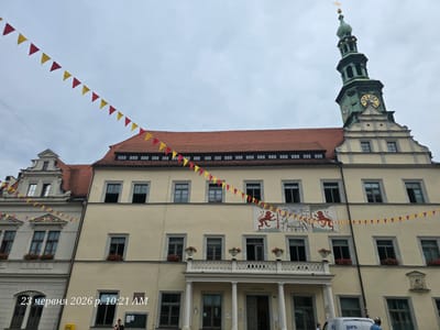 The image shows the exterior of a historic building, likely a town hall, in Pirna, Germany. A tall clock tower with a green spire dominates the right side of the frame. Red and yellow pennant banners are strung across the facade, suggesting a festive occasion. The building has a red tiled roof and light yellow walls, with numerous windows. A balcony with white railings and decorative planters adorns the central section. Above the balcony, a mural depicts heraldic animals, possibly lions, and festive imagery.

In the foreground, a woman is visible near a bulletin board or chalkboard on the building's exterior. Another person can be seen near a white van with the logo "GFG" parked on the right. The sky is overcast, indicating a cloudy day. The bottom left of the image contains text that reads "23 червня 2026 р. 10:21 AM," which translates to "June 23, 2026, 10:21 AM." This suggests the image was taken on that date and time, though the style of the text overlays is typical of a digital timestamp. The overall scene evokes a sense of history and possibly a local celebration or event.