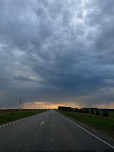 The image shows a long, empty asphalt road stretching into the distance, bordered by green grass and fields on either side. Utility poles with power lines run along the left side of the road. The sky is filled with dramatic, dark grey storm clouds, with a sliver of yellow-orange light visible on the horizon where the sun is setting or breaking through the clouds. The scene appears to be in a rural area. The location is Perevoznoe, Russia. There are no people or animals visible in the image. The weather appears to be overcast and possibly stormy, with rain visible in the distance on the right side of the image.