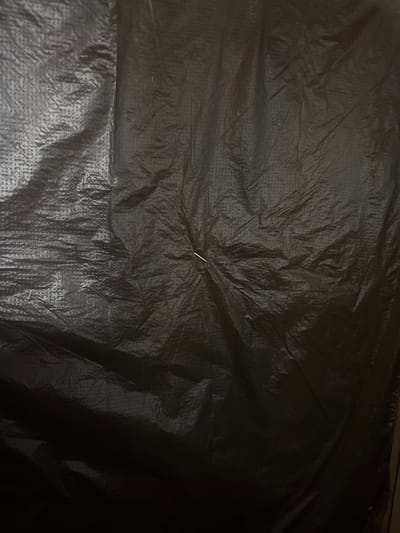 The image is a close-up, full-frame shot of a wrinkled black plastic material, possibly a tarp or large trash bag. The surface has a textured pattern of small, raised dots visible throughout, creating a dimpled effect. The plastic is creased and folded in various directions, with some areas appearing smoother and others more textured due to the light reflecting off the uneven surface. A small, silver metallic object, resembling a staple or a thin pin, is embedded in the plastic near the center of the image. The lighting is dim, casting shadows and highlighting the three-dimensional texture of the material. There is no discernible location in the city of Gribki, Russia, visible in this abstract close-up of a material.