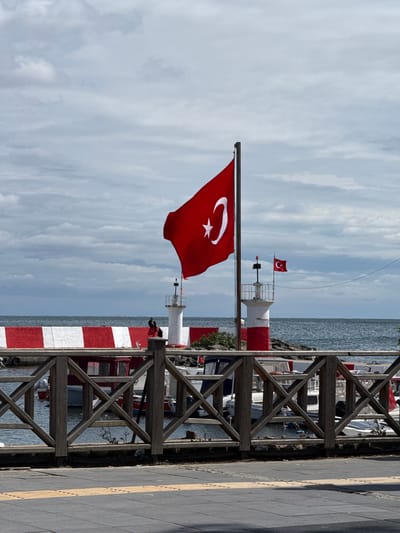 The image depicts a waterfront scene in Tekirdağ, Turkey, under a cloudy sky. A large Turkish flag is prominently displayed on a flagpole in the foreground, fluttering in the wind. Behind it, two lighthouses stand on a pier, one taller and closer than the other. The lighthouses have red and white stripes at their bases. A wooden railing separates the viewer from the water, where several boats are docked. A red and white striped structure, possibly part of a building or barrier, runs along the waterfront. The water appears calm, and the overall atmosphere suggests a coastal town or harbor.