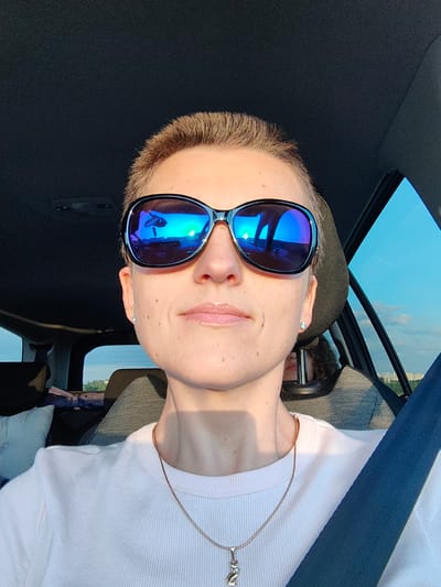 The image is a selfie taken from the perspective of someone sitting in the driver's seat of a car. The subject is a person with short, light brown hair wearing large, dark sunglasses with blue mirrored lenses. The sunglasses reflect the scene outside the car, showing a bright sky with some clouds and a hint of a landscape. The subject is wearing a light pink ribbed t-shirt and a thin gold chain with a pendant shaped like a cat. Their seatbelt is visible across their chest. In the background, another person's head and curly hair can be seen in the passenger seat. The setting appears to be outdoors during daylight, likely late afternoon or early evening due to the warm lighting and long shadows. The window on the right side of the image shows a clear blue sky with some clouds, and a distant city skyline is visible. The overall mood is casual and relaxed. The location is Dzyarzhynsk, Belarus.