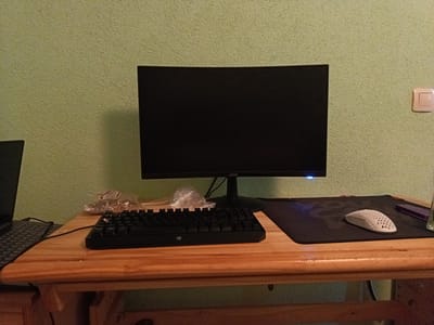 This media file depicts a computer setup on a wooden desk against a textured light green wall. The primary objects are a monitor with the "acer" logo visible, a black keyboard, and a white perforated mouse placed on a black mousepad. A light switch is visible on the wall to the right of the monitor. The scene appears to be indoors, likely in a home or office setting. There is no indication of the time of day, weather, or specific emotional cues. The location is suggested to be Petrich, Bulgaria. The image is static, with no activity or event taking place.