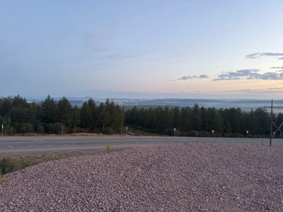 The image depicts a roadside scene at dusk, overlooking a distant city. In the foreground, a large pile of reddish-purple gravel occupies the lower half of the frame. A paved road with white lane markings runs horizontally across the middle of the image, separating the gravel from a dense line of dark green coniferous trees. Beyond the trees, the landscape recedes into rolling hills and a hazy valley. In the far distance, a city skyline is visible, with lights twinkling amidst the fading daylight. The sky above is a gradient of soft blues and pinks, with scattered clouds. The location is identified as Serebryany Bor, Russia. The scene is devoid of any people or visible activity, presenting a quiet, natural landscape with a man-made road and a distant urban settlement.