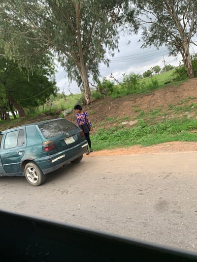 The image depicts a roadside scene, likely in a rural or semi-rural area, under an overcast sky. The photograph is oriented vertically, rotated 90 degrees counter-clockwise.
In the mid-ground, a dark green or blue sedan is parked on the side of an asphalt road, facing away from the viewer. The car shows signs of wear, including white tape on its bodywork near the rear lights. The right rear wheel has a silver five-spoke rim. A license plate is affixed to the rear of the car, but its text is illegible.
Standing near the driver's side rear of the car is a person, dressed in a long-sleeved top with a blue, orange, and black pattern, black trousers or a long skirt, and dark shoes. They also wear a dark head covering or hat. The person is looking towards the left side of the frame, away from the camera.
The road, visible on the right, is made of asphalt with a textured surface. To the left of the road and the car, the terrain consists of a mix of green grass and patches of bare brown earth. Further into the background, beyond a line of fencing or poles, lie expansive green fields dotted with more trees, characteristic of a verdant landscape. Large trees with light-colored bark and abundant foliage dominate the left side of the frame, their branches reaching across the scene. Thin power lines are visible against the cloudy sky above the trees.
The scene suggests daytime under cloudy weather. The setting, with its specific vegetation and road conditions, aligns with the provided location context of Gwaska, Nigeria. No specific activity beyond standing near a parked car is discernible. No legible text is visible other than the unreadable car license plate.