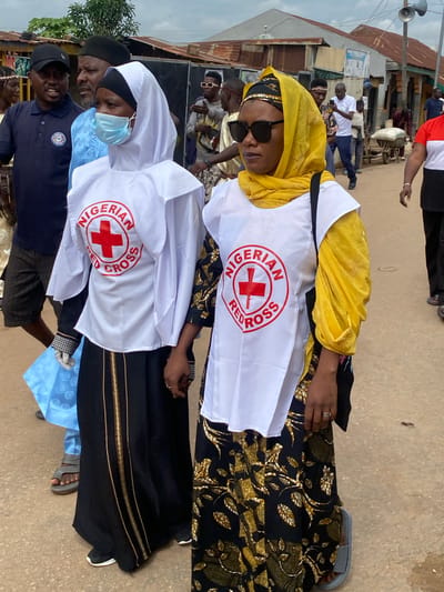 Two women are walking side-by-side outdoors. Both are wearing white vests with the Nigerian Red Cross logo over their clothing. The woman on the left wears a white hijab, a light blue beaded headscarf, and a black skirt with gold trim. She also has a blue face mask on. The woman on the right wears a yellow headscarf and sunglasses. She is wearing a black skirt with a gold leaf pattern. Several other people are visible in the background, including men in traditional clothing and a man in a dark blue polo shirt with a cap. The setting appears to be a street in Rigachikun, Nigeria, with buildings and structures that suggest a market or residential area. The ground is paved with concrete.