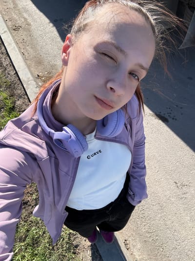 A young person takes a selfie while standing outdoors. The individual has fair skin, one eye winking shut, and is wearing a white t-shirt with the word "CURVE" visible, a lavender jacket, black pants, and purple socks. Headphones of a similar lavender color are draped around their neck. The person is standing on a concrete surface near a patch of grass, likely in an urban setting. The location is possibly in Verkh-Sosnova, Russia.