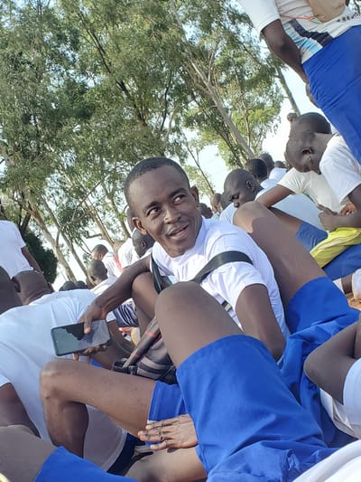 The image shows a group of young men, appearing to be students, gathered outdoors in what seems to be a school or training environment in Barakin Rafin Gora, Nigeria. They are all dressed in similar white t-shirts and blue shorts or trousers. The orientation of the image is sideways.

The individuals are arranged in a cluster, with some seated and others standing. The focus is on a young man in the foreground, who is looking directly at the camera with a slight smile. He is wearing a white t-shirt and black straps, possibly part of a uniform or equipment. He is holding a black smartphone in his right hand.

The setting appears to be outdoors, with trees and foliage visible in the background, suggesting a rural or semi-rural location. The lighting indicates it is daytime, likely with bright sunlight given the clear visibility and shadows. The mood seems relaxed and friendly, with the main subject appearing approachable and engaged.

Some of the other individuals in the group are also visible, with their faces partially obscured or turned away. They share the same attire, suggesting a shared activity or purpose. The overall impression is of a group of young people participating in an outdoor activity, possibly a school event, sports practice, or communal gathering. There is no visible text in the image.