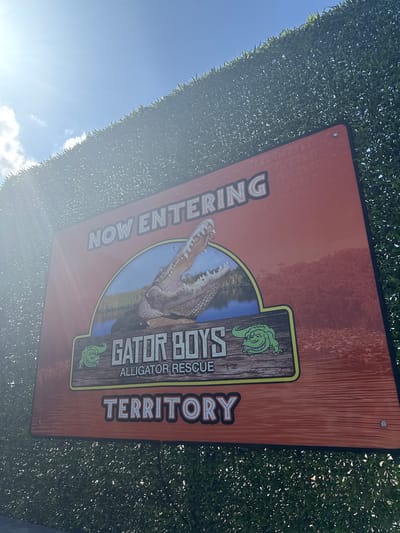 The image depicts a sign that reads "NOW ENTERING GATOR BOYS ALLIGATOR RESCUE TERRITORY." It is attached to a wall of dense greenery and appears to be outdoors, with the sky visible in the background. The sign has an orange background with the text in a bolder font. An alligator is pictured in the center of the sign. Fort Lauderdale, United States, is the location.