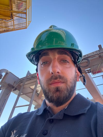 The image shows a man wearing a green hard hat, likely in an industrial setting. A yellow metal structure and a crane are visible in the background, suggesting a construction or industrial site. The location is possibly Marina di Ravenna, Italy, given the prompt's context. The man appears to be looking upwards, perhaps inspecting equipment or structures. He is wearing a dark blue shirt with buttons.
