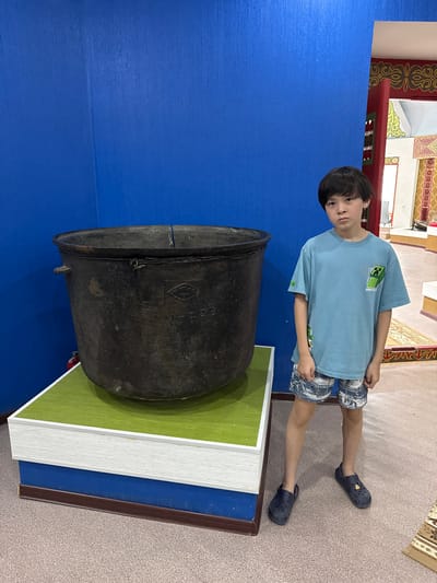 In the city of Fort-Şevtşenko, Kazakhstan, a young boy stands to the right of a large, dark metal pot. The boy is wearing a light blue t-shirt with a Minecraft design and patterned shorts. He is also wearing dark blue clogs. The pot is positioned on a multi-layered display base, with the top layer being a bright green surface. Behind the pot and the boy is a vibrant blue wall. To the right of the blue wall, an opening reveals an interior space with traditional Kazakh decor, including colorful patterns and intricate designs. The floor is carpeted with a light brown pattern. The overall environment suggests a museum or cultural exhibition.