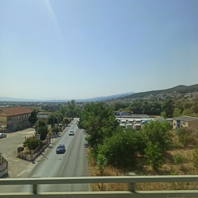 The image depicts an elevated view of a road in the city of Lagyna, Greece, on a clear, sunny day. The road runs from the foreground to the middle ground, curving slightly to the right. Two vehicles are visible on the road: a dark-colored SUV in the right lane, and a lighter-colored sedan further ahead in the left lane.
To the left of the road, there is a building with a red-tiled roof and several dark-tinted windows, suggesting it might be a commercial establishment. Adjacent to this building, a lighter-colored car is parked. A metal fence with a gate separates the sidewalk from a grassy area.
On the right side of the road, there are several mature trees and dense vegetation. Beyond the trees, a collection of white trailers or mobile homes are parked in an open lot. Further in the background, a hilly landscape is visible, dotted with buildings, indicating a town or city extending up the slopes. The sky is a clear, pale blue, with a subtle light flare in the upper left quadrant. A light grey metal railing runs across the bottom of the image, suggesting the viewpoint is from a bridge or elevated walkway.