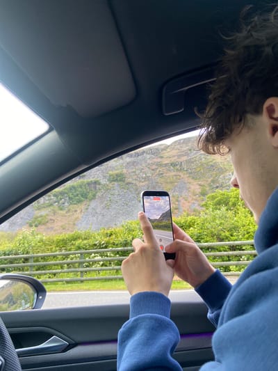 From inside a car, a person wearing a blue sweatshirt is filming a rocky hillside and a road in Llangynog, United Kingdom. The phone screen reflects the scene. The car interior is visible. The window is slightly down.