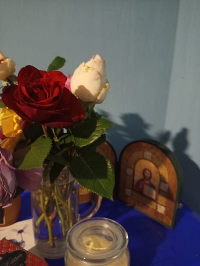 The image depicts a still life arrangement on a blue surface, likely a table or shelf, against a light blue wall. In the foreground, a clear glass vase holds a bouquet of roses in various stages of bloom and color, including red, white, yellow, and purple. Some roses are fully open, while others are still buds. Green leaves are interspersed among the flowers. To the right of the vase, a small, arched wooden icon is visible, featuring a religious depiction, likely of Jesus Christ. The icon casts a shadow on the wall behind it. In the lower right corner, a clear glass jar with a lid is partially visible. In the lower left, a red and gold patterned box and a white card with a graphic of a person riding a bicycle are also present. The lighting suggests an indoor setting, possibly with natural light casting shadows. The overall scene conveys a sense of domestic tranquility and devotional practice. There is no indication of the city of Чэрвеньскі сельскі Савет, Belarus within the image itself.