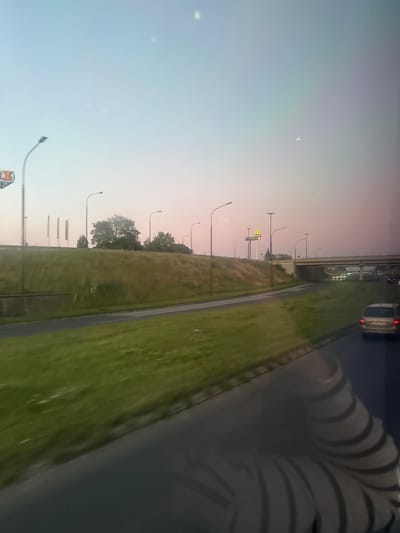 The image depicts a road scene at dusk or dawn, with a soft pink and blue gradient in the sky. In the foreground, a dark, reflective surface, likely the hood or windshield of a vehicle, shows a striped pattern of light and shadow. A gray car is visible on the right side of the road. The road curves to the left and is bordered by a grassy embankment.

Further in the background, a McDonald's sign with its iconic golden arches is illuminated. Several streetlights are positioned along the road and embankment. An overpass stretches across the scene, with cars visible beneath it. The area appears to be an urban or suburban environment, potentially an approach to or exit from the city of Lublin, Poland, given the presence of the McDonald's and highway infrastructure. The overall atmosphere is serene and quiet.