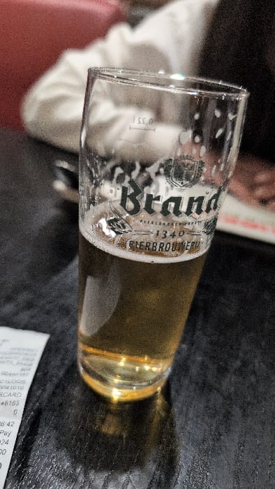 The image displays a clear glass of "Brand" beer, approximately one-third full with golden-amber liquid and residual foam, situated on a dark, textured tabletop. The glass features the "Brand" logo, "KLEINBLUSCH - 1340 - BIERBROUWERIJ," and a "0,5 L" fill line. To the left, a white paper receipt is partially visible, showing text fragments including "LOGS," "CARD," "*8153," "26:42," and "Pay." In the blurred background, a person wearing a light-colored, long-sleeved garment is partially discernible, seated near a reddish upholstered surface. Other indistinct forms suggest a social setting. The presence of Brand beer, a Dutch brewery, places the scene within the city of Horst, Netherlands, likely in a commercial establishment such as a restaurant or bar.