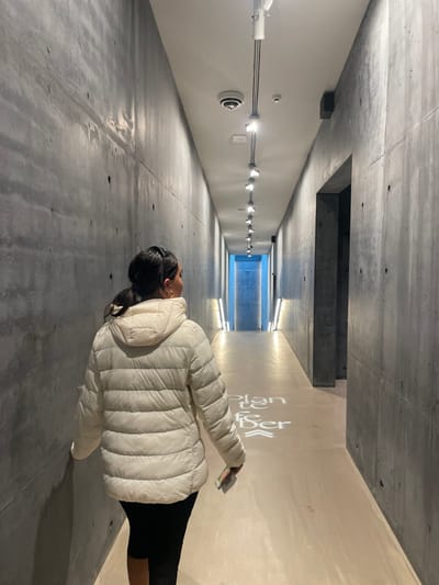 A woman is depicted walking away from the camera down a minimalist concrete corridor. She is positioned slightly right of center, her back to the viewer, and her left shoulder turned slightly forward. She wears an off-white quilted puffer jacket with a hood and dark-colored leggings. Her dark hair is pulled back, secured by a white scrunchie. A small gold hoop earring is visible on her left ear. Her left hand is near the concrete wall. In her right hand, she holds a small, rectangular object, possibly a phone or card, displaying a partial green color.

The corridor is characterized by raw, exposed concrete walls with visible formwork lines and tie holes. The ceiling is white, featuring a track lighting system with multiple individual spotlights illuminating the path. A white dome-shaped security camera and a small red indicator are mounted on the ceiling in the foreground. The floor is a light-colored, smooth surface. White text, partially visible as "Plan to ser" with two upward arrows, is projected onto the floor as directional signage further down the hallway. Approximately midway down the right wall, a dark, rectangular entryway or recess is visible. The far end of the corridor opens into an area intensely illuminated by blue light, flanked by two vertical, bright light fixtures. The scene is located in a modern architectural setting within Klampenborg, Denmark.