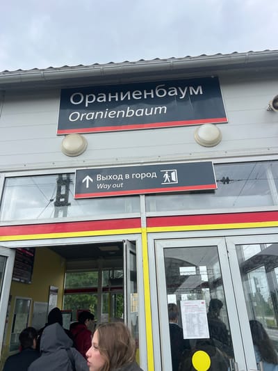 The image shows the exterior of a train station building in Lomonosov, Russia. Above the entrance, a dark sign with white text reads "Ораниенбаум" and below it, "Oranienbaum." To the right of this main sign, a smaller dark sign with a white arrow pointing up and an icon of a person exiting a door indicates "Выход в город" (Exit to the city) and "Way out." The building's facade is grey, with red and yellow trim above the entrance. Through the glass doors and windows of the entrance, several people are visible queuing inside. The sky is overcast.