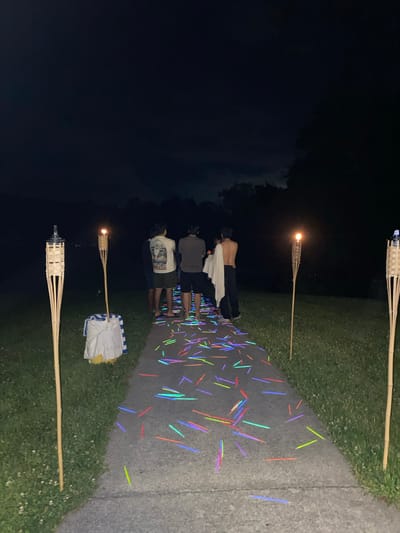 The image depicts an outdoor nighttime scene, likely a pathway or walkway. The ground is illuminated by numerous glow sticks scattered across it in various colors, including pink, blue, green, and orange. Two tiki torches stand on either side of the walkway, one on the left and another on the right, providing ambient light. In the background, a group of four individuals are visible from behind, standing on the path. The environment appears to be a grassy area bordering the walkway, with trees silhouetted against the dark night sky. The location is specified as Washington Township, United States of America.