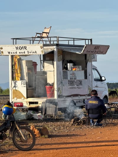 The image depicts a food truck named "NOIR Wood and Drink" and "NOIR Food and Drink Truck" parked on a dirt road in Trường Thọ, Vietnam. A security guard, wearing a dark blue jacket with a yellow "SECURITY" patch on the back, sits in a black folding chair facing away from the camera. In front of the security guard, a small fire burns, producing smoke. Adjacent to the food truck is a motorcycle with a blue and yellow helmet. A black dog is visible in the background, near a table and chairs set up outdoors. The food truck itself is white with colorful decals. One decal advertises "FORNO DI LEGNA - WOOD-FIRED PIZZA." Above the truck, a light pink folding chair is placed on a raised platform. The overall setting appears to be an open area with sparse vegetation and distant hills under a clear blue sky.