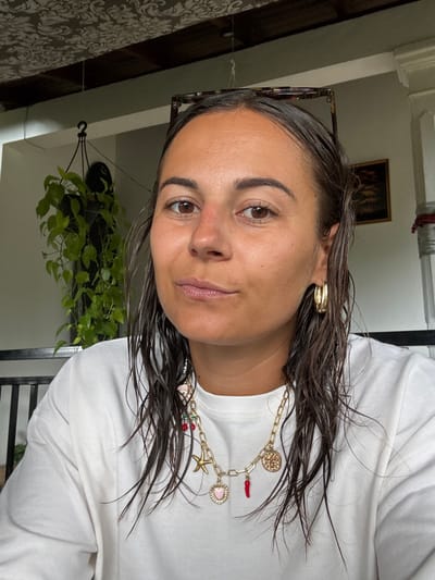 An individual, identified as female, is depicted in a close-up, front-facing portrait, likely a selfie, within a location in Nilwella, Sri Lanka. She has tanned skin, dark brown wet hair, appearing medium-length and falling over her shoulders. Her expression is neutral to slightly smiling, with brown eyes and subtle freckles visible on her nose. She wears a plain white crew-neck long-sleeve sweatshirt. Accessories include gold-colored hoop earrings in both ears and a layered gold-colored chain necklace adorned with multiple charms, specifically a red chili pepper, a pink heart, a gold starfish, and a circular gold medallion. Dark-framed tortoiseshell sunglasses are resting on top of her head.
The background indicates an indoor or covered outdoor setting. The ceiling features dark wooden beams overlaid with a light-colored, ornate damask-patterned material. To the left, a hanging plant with trailing green leaves is suspended in a dark pot from the ceiling by a rope and hook. White wall or pillar sections are visible, and behind the subject's left shoulder, a dark-colored railing or furniture piece is partially present. On the far right, a framed picture with dark, possibly landscape, imagery hangs on a wall, and a red and white striped element is visible above it. Natural light illuminates the scene from an unspecified source.