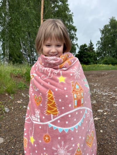 A young fair-skinned child with short, light brown hair and a fringe is positioned centrally in an outdoor setting, identified as Kryukovo, Russia. The child, approximately three to six years old, is smiling with a downward gaze, revealing teeth. She is completely enveloped in a plush, pink blanket adorned with various Christmas-themed motifs. Visible patterns on the blanket include white snowflakes, orange and yellow Christmas trees, brown gingerbread houses with blue and red accents, yellow stars, white gift boxes, red and orange circular ornaments, and white script that partially reads "Merry". A string of blue and white triangular bunting is also depicted on the blanket.

The background consists of a natural environment under an overcast sky. Behind the child, deciduous trees with green foliage and light-colored trunks, possibly birch, are visible, along with a darker coniferous tree trunk on the right. Further back, denser green foliage and trees extend across the horizon line. The ground beneath the child and extending into the foreground is a mix of small gravel, dirt, and sparse green vegetation. Patches of taller green grass and weeds are present on the left side of the frame. The overall impression is a somewhat muted, natural outdoor scene on a cloudy day.