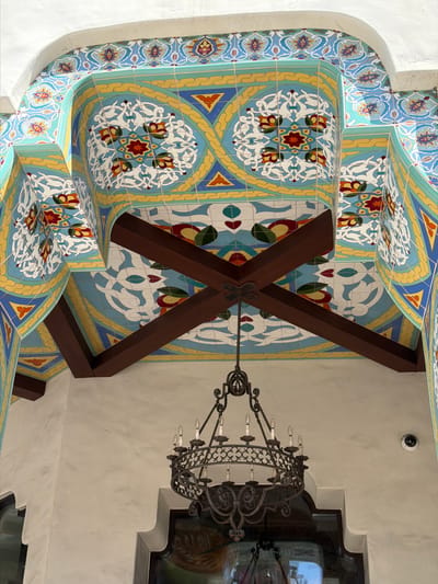 A low-angle, eye-level shot captures the intricately decorated interior archway and ceiling of a building. The arched ceiling is adorned with vibrant, geometric tiles featuring floral motifs in shades of blue, yellow, red, and white. Two dark wooden beams cross diagonally overhead, supporting the ceiling. Hanging from the center of the beams is an ornate, black wrought-iron chandelier with multiple candle-style lights. The walls are a neutral, textured cream color. A security camera is visible on the right wall. The scene is located in Pasadena, California, United States of America, and appears to be an entrance or covered walkway. There are no people present in the image.