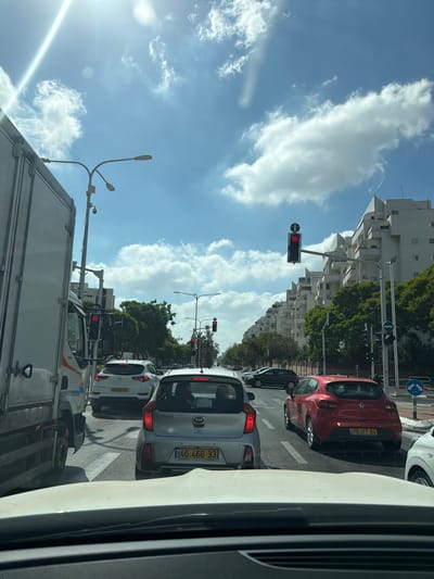 This is a vertical, medium-angle, eye-level shot of traffic in Ashdod, Israel, on a bright, sunny day with scattered clouds. The image is dominated by a busy intersection with several cars stopped at a red light. In the foreground, the hood of a white car is visible.
Immediately behind the white car is a silver Kia Picanto with license plate "46-466-33". To its right is a red Renault Clio with license plate "75-177-84". Further back, a white Hyundai Tucson with license plate "69-081-55" is also stopped. A large white truck occupies the left side of the frame, obscuring some of the view of the intersection.
Multiple traffic lights are visible, all showing red, indicating the traffic is stopped. Streetlights and utility poles are present, one with a security camera attached. Tall, white apartment buildings with distinctive architectural features line the right side of the street, interspersed with lush green trees. The sky is a clear blue with fluffy white clouds, and the sun is bright, casting a strong glare in the upper left corner of the image. The scene depicts a typical urban traffic situation on a clear day.