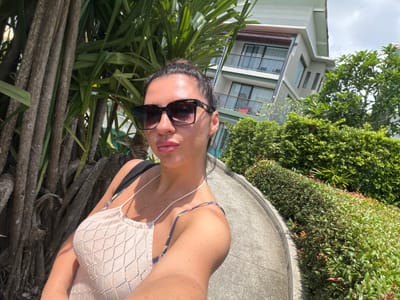 A woman takes a selfie outdoors in a tropical setting. She is wearing large, dark sunglasses with a light-colored, open-knit top. Her hair is pulled back into a bun, and she is wearing a delicate necklace. In the background, a modern building with balconies is visible, surrounded by lush green trees and bushes. The path in front of her is paved with a light-colored, curved material. The overall scene suggests a warm, sunny day.