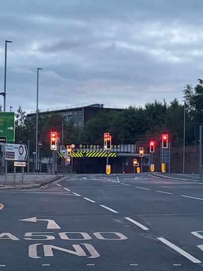 This vertically oriented photograph captures a complex road junction in Stoke-on-Trent, United Kingdom, under an overcast sky. The scene is viewed from a stationary vehicle, facing forward towards an array of traffic signals and a bridge structure.
In the foreground, the dark asphalt road is marked with white dashed and solid lines, a yellow box junction, and painted road text including the partial word "TURN" and "A500" with an arrow, indicating a turn lane.
The mid-ground is dominated by numerous traffic lights mounted on poles and gantries. Most display red lights, indicating that vehicles are stopped. Several amber/yellow signals are also active, including arrow indicators for various turning movements.
To the left of the road, a large green directional sign points straight ahead, listing "City Centre" and "A52 (Hanley)". Below this, a white sign indicates "City entry", "A5006", "Cultural Quarter", and "5 mi. Longton". Further along, a prominent yellow and black striped barrier or structure, likely part of a bridge or overhead clearance, clearly displays the text "Bridge head". Below this, a red circular sign shows a height restriction of "4.0m" (13'-0"), and another red circular sign with a white bar (no entry) is visible.
In the background, beyond the road infrastructure, dense green foliage and a multi-story brick building with several windows are visible. The sky is a uniform light grey, characteristic of a cloudy day. The overall scene depicts a busy urban intersection, with numerous signals and signs guiding traffic.