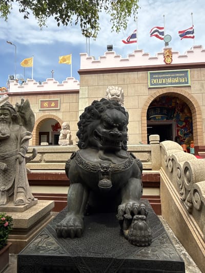 The image shows the exterior of a temple in the city of Mab Frak Thong, Thailand. In the foreground, a large stone lion statue sits on a dark pedestal. Behind the lion, a statue of a warrior figure stands to the left, and the entrance to the temple, an arched doorway decorated with colorful murals of dragons, is visible. Above the entrance, a sign with Thai script is mounted on the stone wall. Thai flags and yellow flags with a circular emblem fly on poles atop the temple's crenellated facade. The sky above is partly cloudy.
