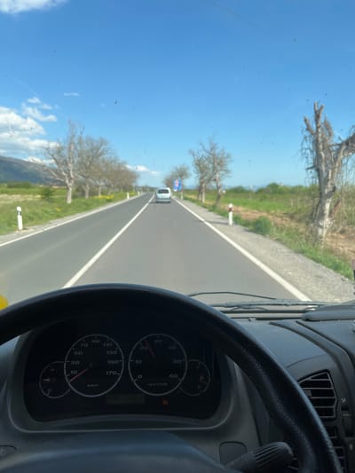 The image shows the view from the driver's seat of a vehicle moving along a road on a sunny day. The road is straight and appears to be paved, with trees lining the side. Another vehicle is visible in the distance. The dashboard of the vehicle is visible, including the steering wheel and speedometer. The speedometer indicates a speed of approximately 10 km/h. The sky is blue with some clouds. There is no information in the image to indicate the location in Pevtsite, Bulgaria.