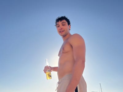 A man is holding a bottle of Corona beer against a clear blue sky. He is shirtless, wearing light-colored swim trunks with a drawstring. His hair is dark and curly, and he has a smiling expression. The sun is bright, creating a backlight that highlights the man and the beer bottle. The scene appears to be outdoors, possibly at a beach or on a boat, given the presence of a distant sailboat mast in the lower right corner of the image, consistent with a Mediterranean location. The lighting suggests it is daytime, likely late afternoon or early evening given the angle of the sun. The overall mood is relaxed and celebratory. The visible text on the beer bottle reads "Corona Extra".