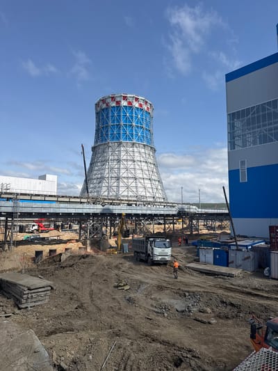 The image depicts a construction site in Chulman, Russia, dominated by a large, unfinished cooling tower with a white lattice structure and blue and red accents at the top. Adjacent to the cooling tower is a partially constructed industrial building with a blue and grey facade. The foreground is a muddy construction area with piles of dirt, concrete slabs, and construction equipment, including a white truck, a yellow excavator, and a red truck. Several workers in orange vests are visible, some operating machinery and others walking. In the background, there are more industrial structures and a treeline under a cloudy blue sky. The scene is one of active construction and industrial development.