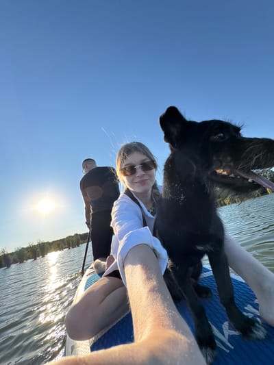 This is a selfie taken from a paddleboard on a body of water under a clear blue sky. The sun is setting or rising, casting a bright glare on the water and behind a person standing on the paddleboard. A woman is in the foreground, holding the camera and smiling. Next to her is a black dog, looking towards the camera with its mouth open. Another person is standing on the paddleboard in the background, holding a paddle. The paddleboard is blue and white. Trees are visible in the distance along the shoreline. The location is identified as Novotitarovskaya, Russia.