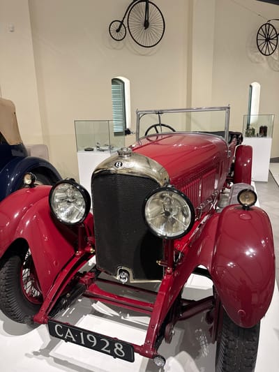 The image shows a red vintage car, specifically a 1928 Bentley, displayed in what appears to be a museum or exhibition hall in Stellenbosch Local Municipality, South Africa. The car has a black grill with the Bentley logo prominently displayed, large headlights, and a license plate that reads "CA-1928." The background is a neutral off-white color, with various artifacts such as bicycles displayed on the wall.