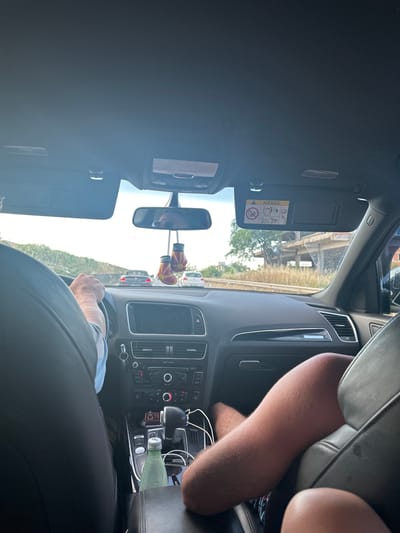 The view from inside a moving car shows the driver's hands on the steering wheel. Dangling from the rearview mirror are boxing gloves with the word "Macedonia" on them. The passenger has a leg exposed in the back seat. A water bottle is visible on the center console. The sun visor is down. The location outside the car appears to be on a highway, likely in Καρδία, Greece. Other cars are visible in the distance.