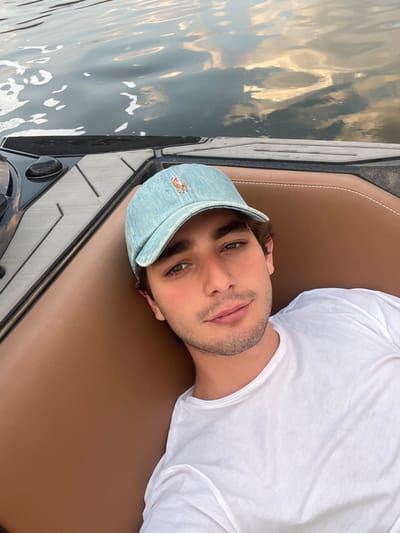 A man with olive skin and brown hair is laying down in a boat. He is wearing a light blue baseball cap and a white t-shirt. He is looking at the camera with a slight smile. The boat has a brown leather seat, a black console, and a window showing the water. The water reflects the sky and appears to be calm.