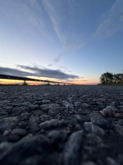 A low-angle, ground-level shot captures a gravel surface in the foreground, with small, dark gray stones filling the frame. Beyond the gravel, the image transitions to a blurred background depicting a road or path leading towards a horizon line. A dark, elevated structure, possibly a bridge or overpass, stretches horizontally across the middle ground, illuminated by a faint orange glow from the setting sun. The sky above is a gradient of pale blue at the top, fading to a soft orange and pink near the horizon, with wispy clouds present. To the right of the frame, a line of trees is visible in the distance, their silhouettes dark against the colorful sky. The scene appears to be an outdoor location in Paldiski, Estonia, likely near a road or thoroughfare during dusk. No people or specific actions are discernible in the image.