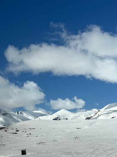 The image depicts a bright, clear day in Ganisi, Georgia, likely in a mountainous region. A crisp blue sky is partially obscured by thick, puffy white clouds. Adjacent to the sky, a snow-covered mountain range stretches into the distance. The foreground shows a snow-covered terrain with sparse infrastructure elements, such as small rectangular structures, possibly ski equipment or buildings. The scene is mostly devoid of human presence.