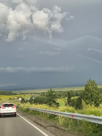 The image shows a white Honda CR-V driving on a paved road in a rural area, possibly Verkh-Chita, Russia, given the license plate. The car is in the foreground, on the left side of the road, facing away from the camera. The road curves to the right and is bordered by a metal guardrail with red reflectors. Lush green vegetation, including tall grass and trees, grows on either side of the road. In the background, there is a vast landscape of rolling hills covered in fields and scattered trees. The sky is overcast with dark, heavy clouds, suggesting an approaching storm. There are no people visible in the image. The overall scene depicts a scenic drive through a countryside under a dramatic sky.
