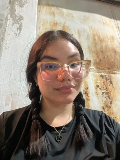 A young woman with dark hair in two braids and wearing glasses with light-colored frames and a black t-shirt with a heart pendant is pictured from the chest up. She is in front of a textured wall. The lighting suggests it is evening. There is no information about the location of Conuco Viejo, Venezuela.