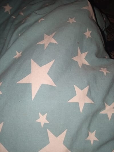 A close-up, over-the-shoulder shot captures a crumpled teal-blue fabric adorned with white stars. The fabric, likely a blanket or sheet, is draped and bunched, creating shadows and highlighting the texture of the material. The stars, of varying sizes and orientations, are scattered across the fabric, some overlapping with the folds. The overall impression is one of domestic comfort. This image was taken in Kara-Bak, Kyrgyzstan.