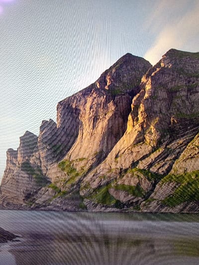 The image depicts a natural landscape situated in Olambe, Nigeria. Dominating the scene are massive, rugged mountains or cliffs that ascend directly from a calm body of water, which occupies the lower portion of the frame. The rock surfaces display varying shades of grey and brown, featuring prominent vertical and diagonal stratification, with interspersed patches of green vegetation, indicative of moss or sparse plant growth, on the slopes. The upper sections of the mountains culminate against a light, pale blue sky that exhibits faint cloud formations towards the right. Portions of the rock faces are illuminated by a warm, golden light, creating distinct contrasts with shadowed areas, suggesting the scene is captured during sunrise or sunset. The surface of the water is a muted grey-blue, showing subtle ripples and reflections of the overlying sky and adjacent mountains. There are no visible people, animals, man-made structures, actions, or interactions within the scene. A noticeable diagonal moiré pattern is present across the entire image, consistent with a photograph taken of a digital screen.