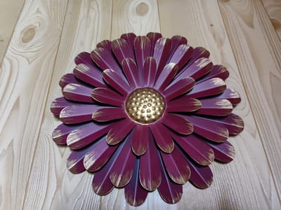 This media file features a close-up, top-down view of a decorative metal flower with deep burgundy petals accented with gold at their tips. The flower is positioned on a light-colored wooden surface, likely a tabletop or wall panel, with visible wood grain patterns. The setting appears to be indoors, with no indication of time of day or weather conditions. The flower is the sole subject, with no people or other objects present in the frame. The lighting is even, suggesting a well-lit indoor environment. The overall impression is one of decorative artistry and simple, clean presentation. The location context provided is Rybnitsa, Moldova, but there are no specific visual cues within the image to confirm this location. There is no visible text in the image.