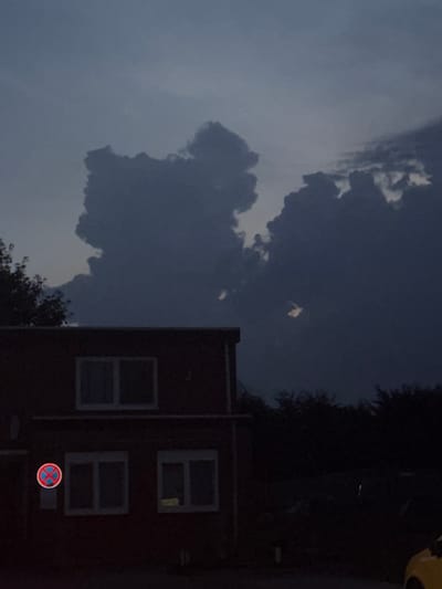 The image shows a building in Neumünster, Germany, under a dark, cloudy sky. The building appears to be a two-story structure made of brick. There are several windows visible on the facade, some of which are illuminated. A red and blue circular traffic sign with an "X" shape is mounted on the wall near the ground floor. The sky is filled with large, dark clouds that are illuminated from behind, creating a dramatic effect. The surrounding area is dark and appears to be an outdoor space with trees and vegetation. The overall mood of the image is somber and somewhat ominous, likely due to the dark clouds and the late hour.
